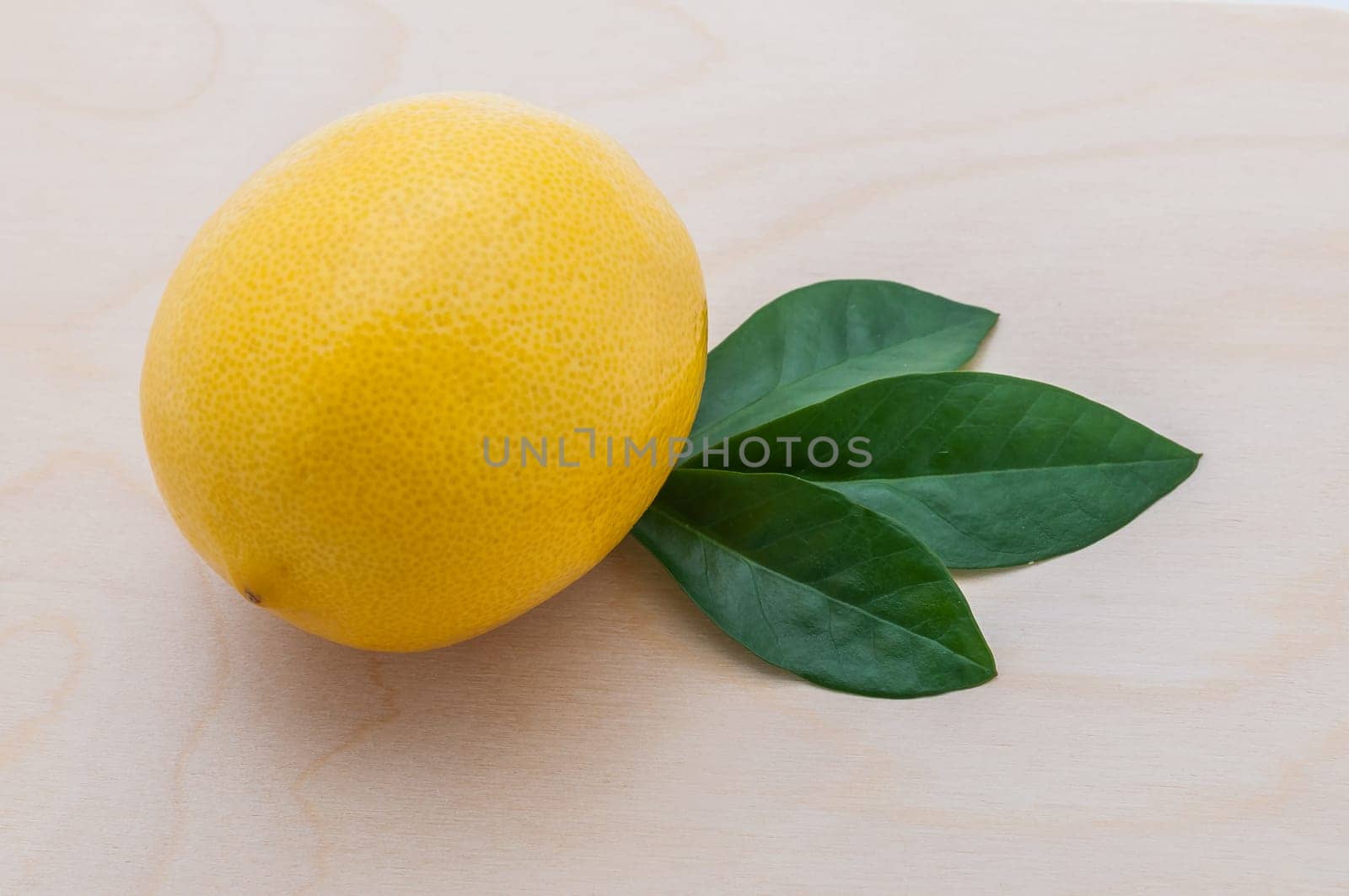 close-up of a yellow bright lemon with three green leaves on a chopping board of light wood.