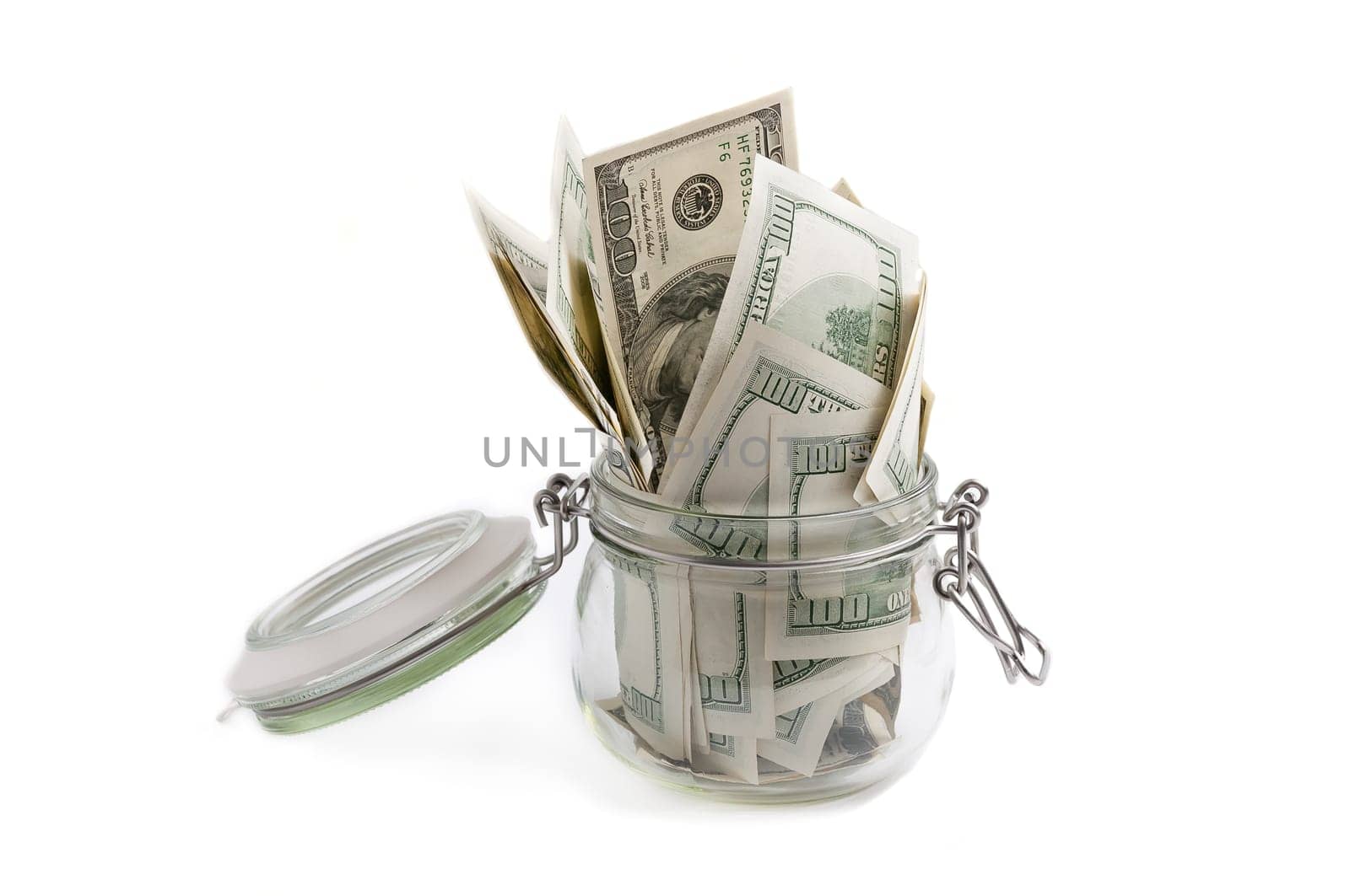 A glass jar filled with dollars among a hundred dollar bills on a white background.