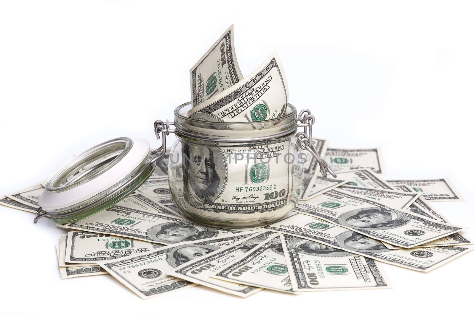 A glass jar filled with dollars among a hundred dollar bills on a white background.