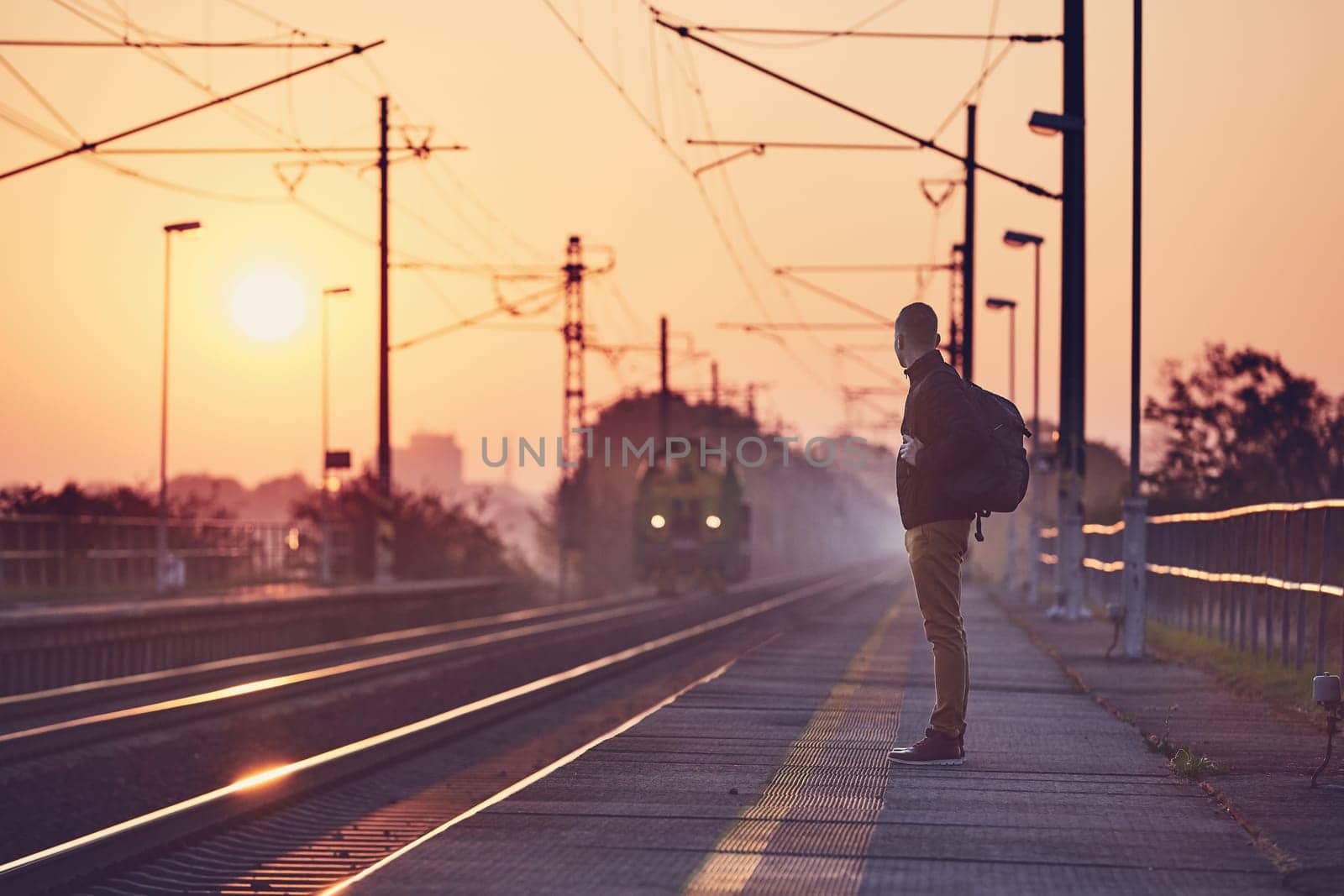 Alone man with backpack waiting at railroad station platform at sunrise.