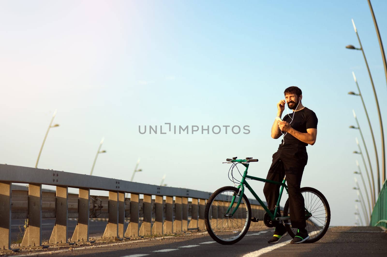 The smiling cyclist is using phone with earphones for listening music outdoors on the bridge.
