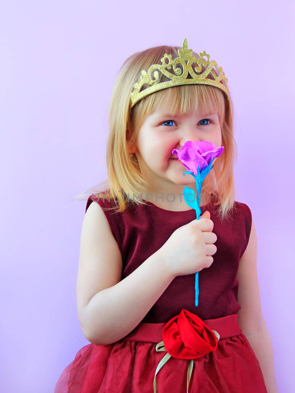 Little girl dressed as princess with crown smelling artificial flower. Little Princess. Girl smelling flower and smiling. Child in crown. Portrait of girl princess