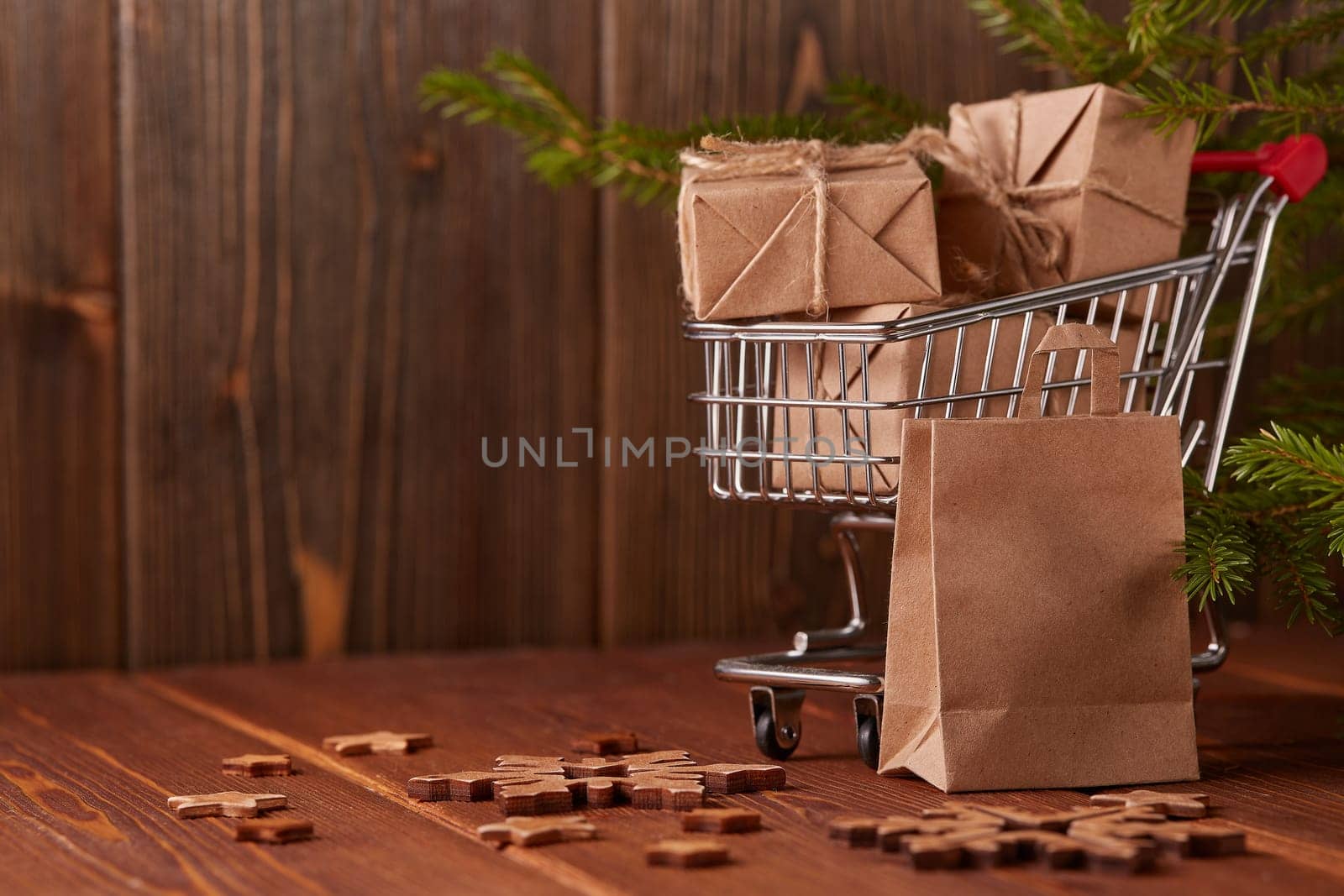 Shopping trolley with gift boxes and fir tree branches on a brown wooden background. Christmas and New Year shopping.