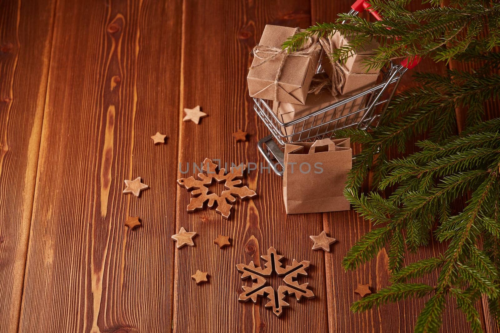 Shopping trolley with gift boxes and fir tree branches on a brown wooden background. Christmas and New Year shopping.
