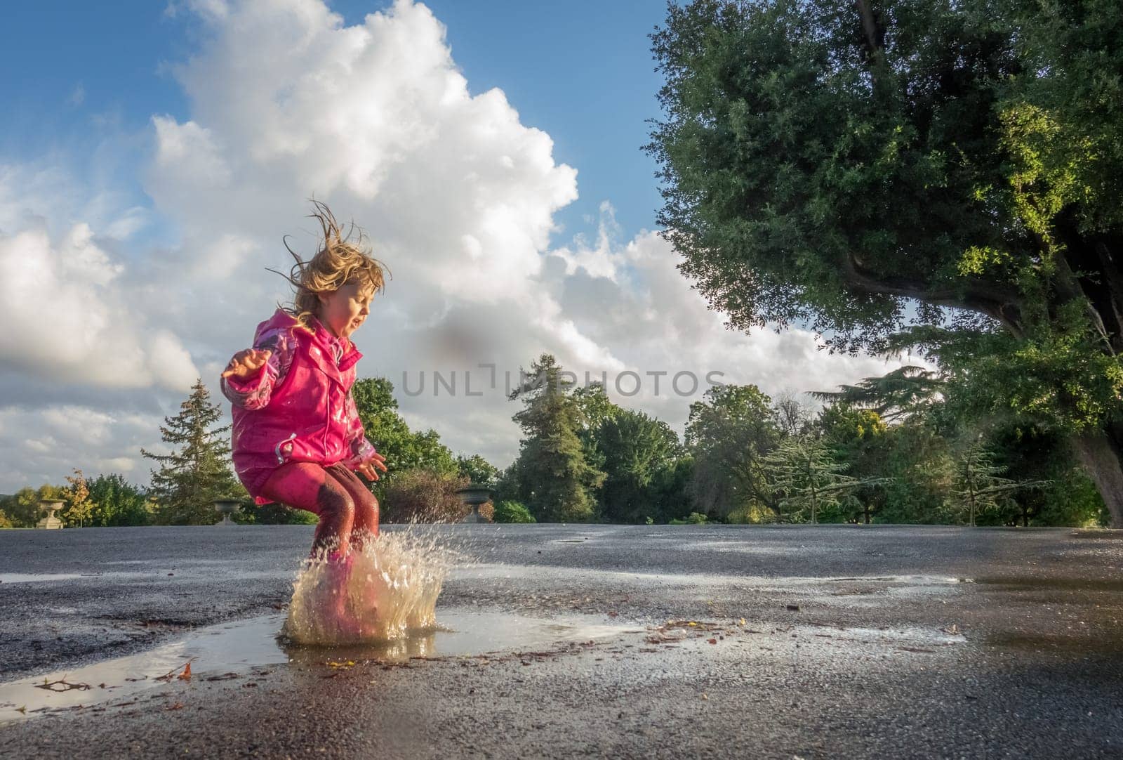 Little Caucasian girl jumping into the puddle after rain in english countryside