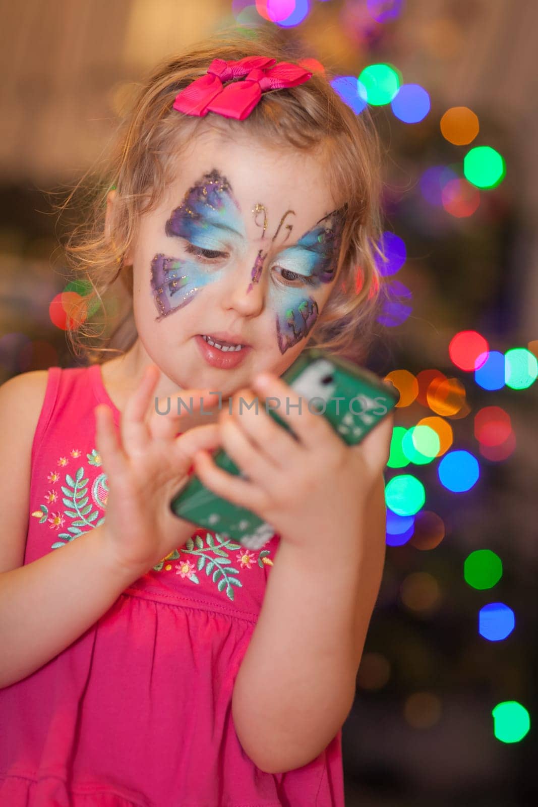 Cute little Caucasian girl having butterfly painted on her face during children birthday party, playing with her moms smartphone
