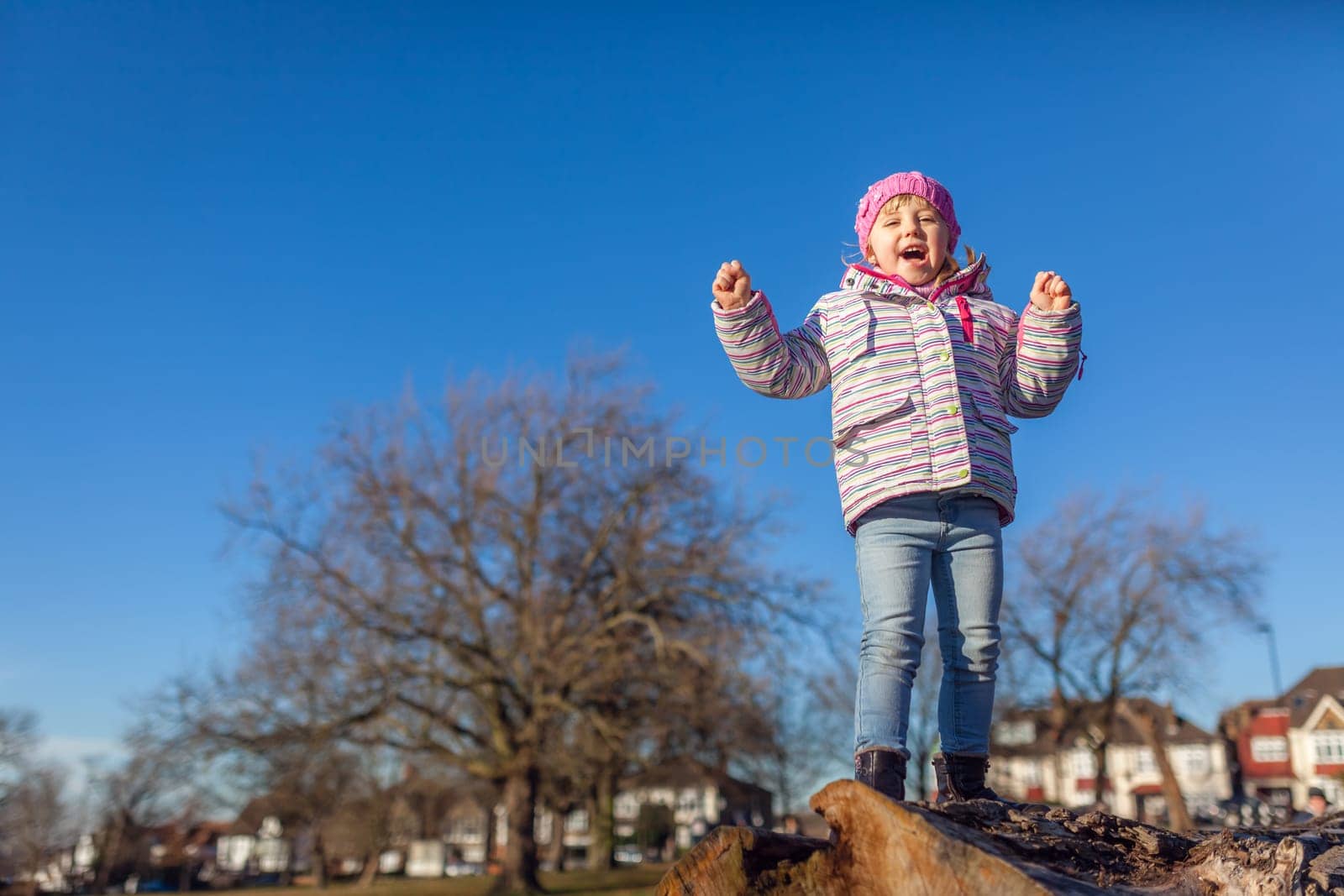 Cute little girl shouting from joy while standing on the top of large cut tree log in the park in spring