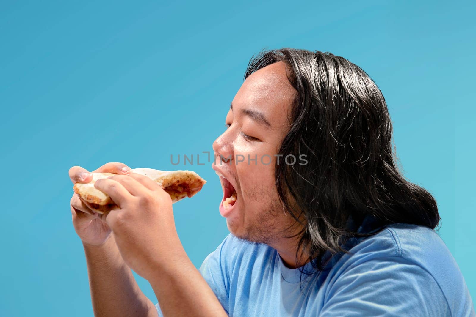 Asian fat man eating slice of pizza fast food over colored background