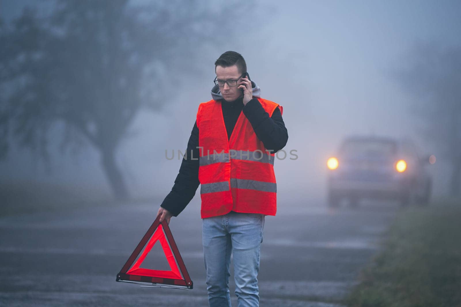 Traffic problem in thick fog. Young driver calling for assistance and putting warning triangle behind his broken car.