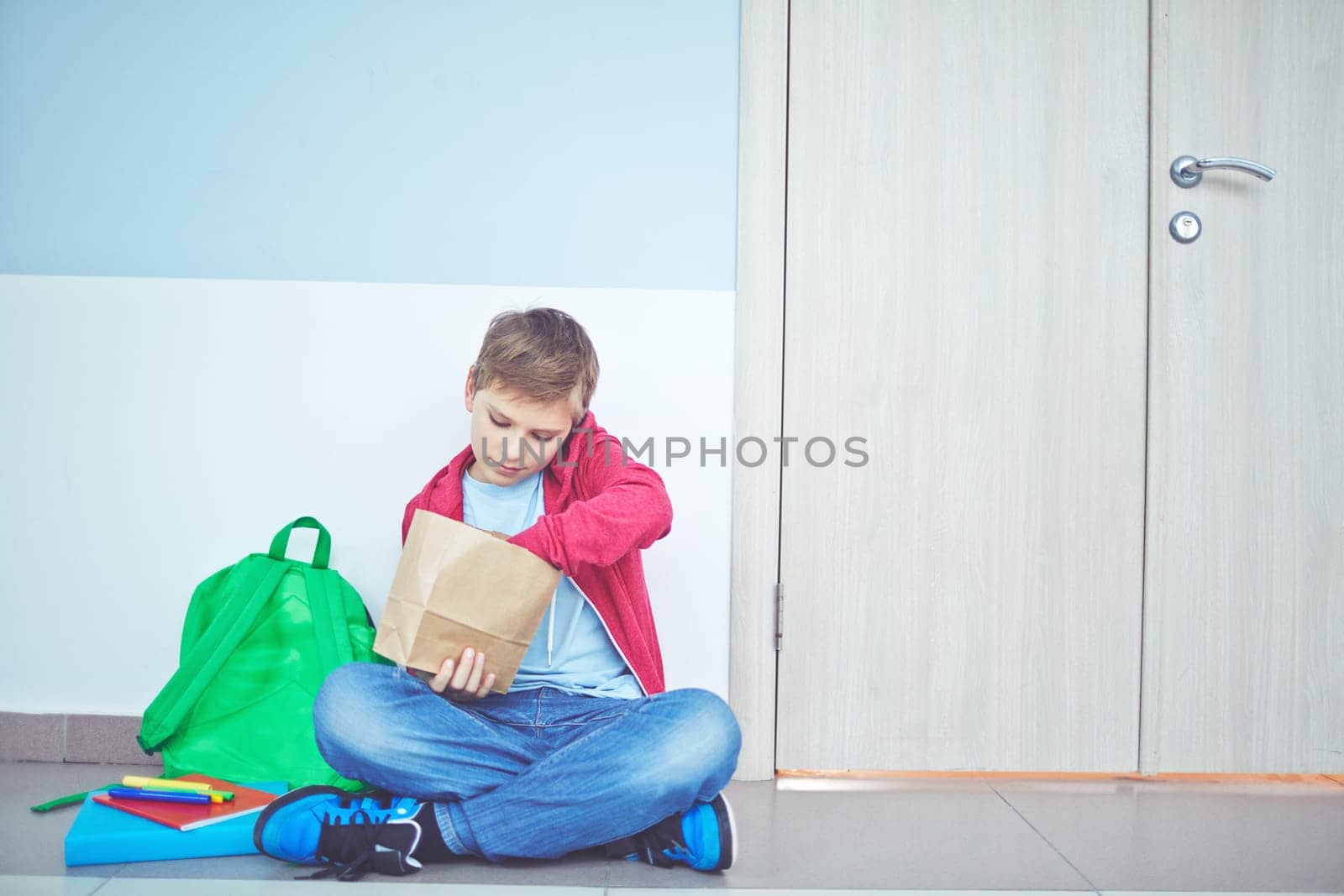Cute schoolboy unpacking his lunch while sitting on the floor by classroom door