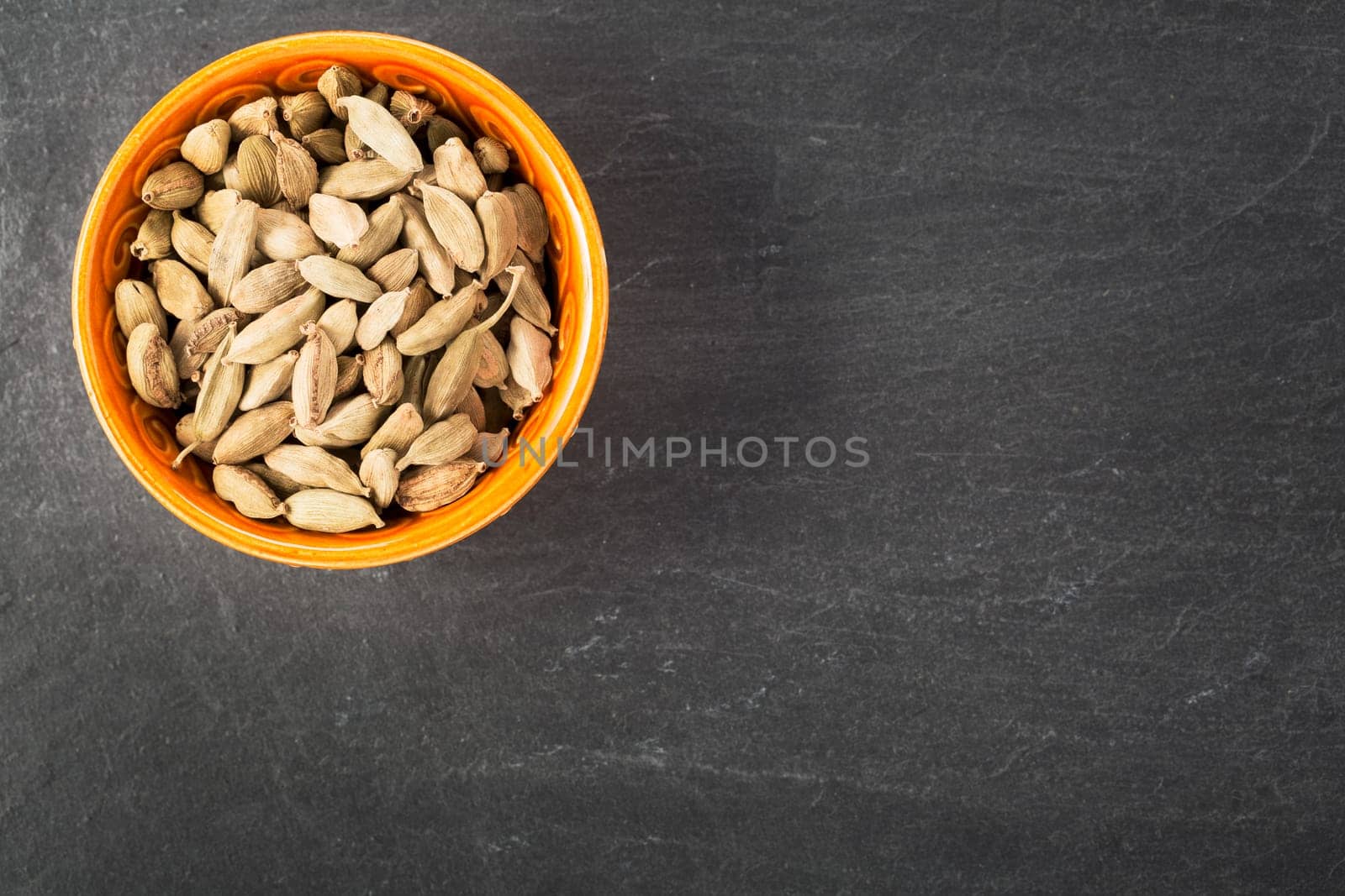Whole Cardamom seeds in orange bowl on grey stone surface with copy space.