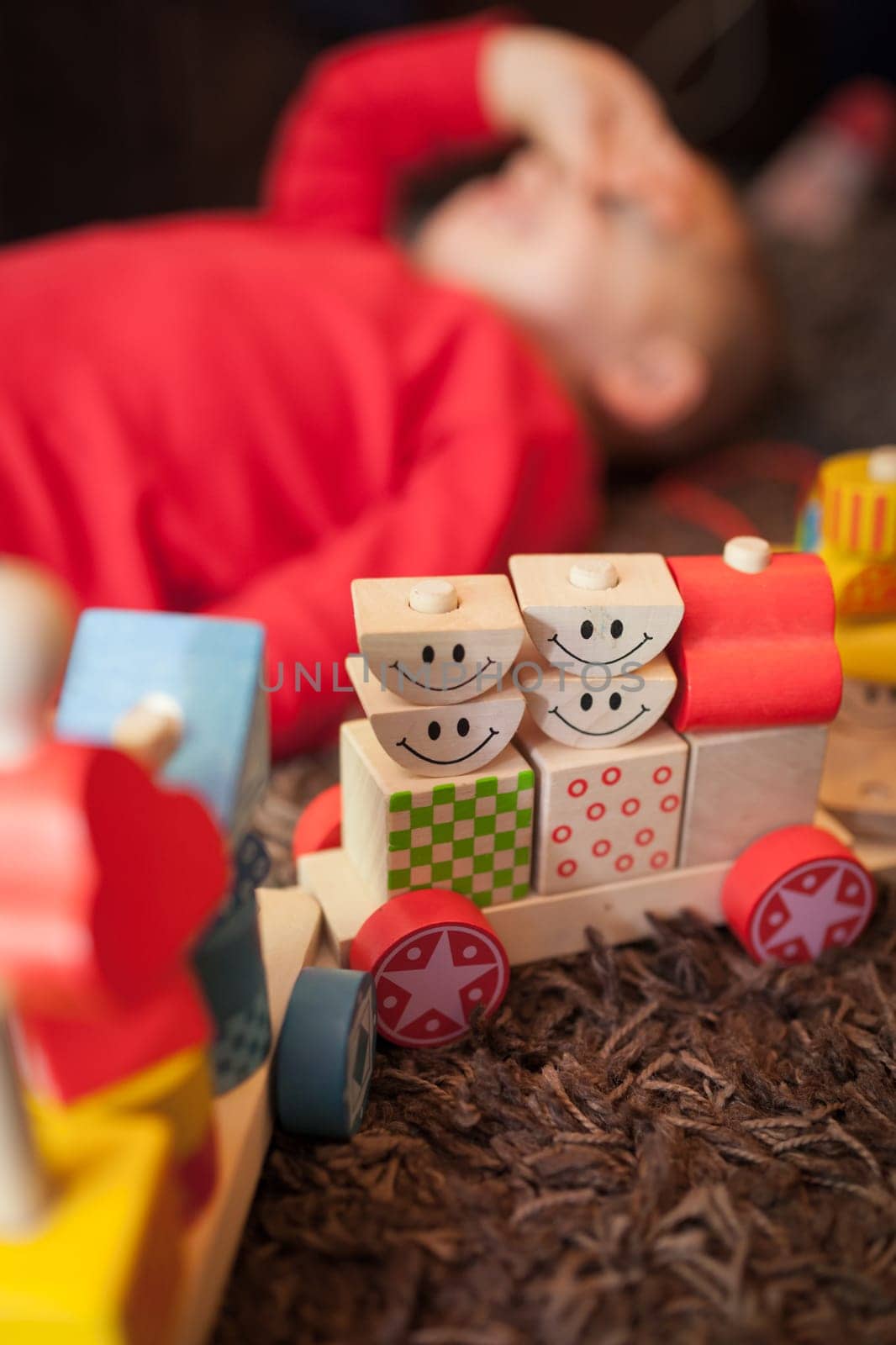 Little Caucasian boy lying tired after playing with a colourful wooden toy train on the carpet in the living room in the house
