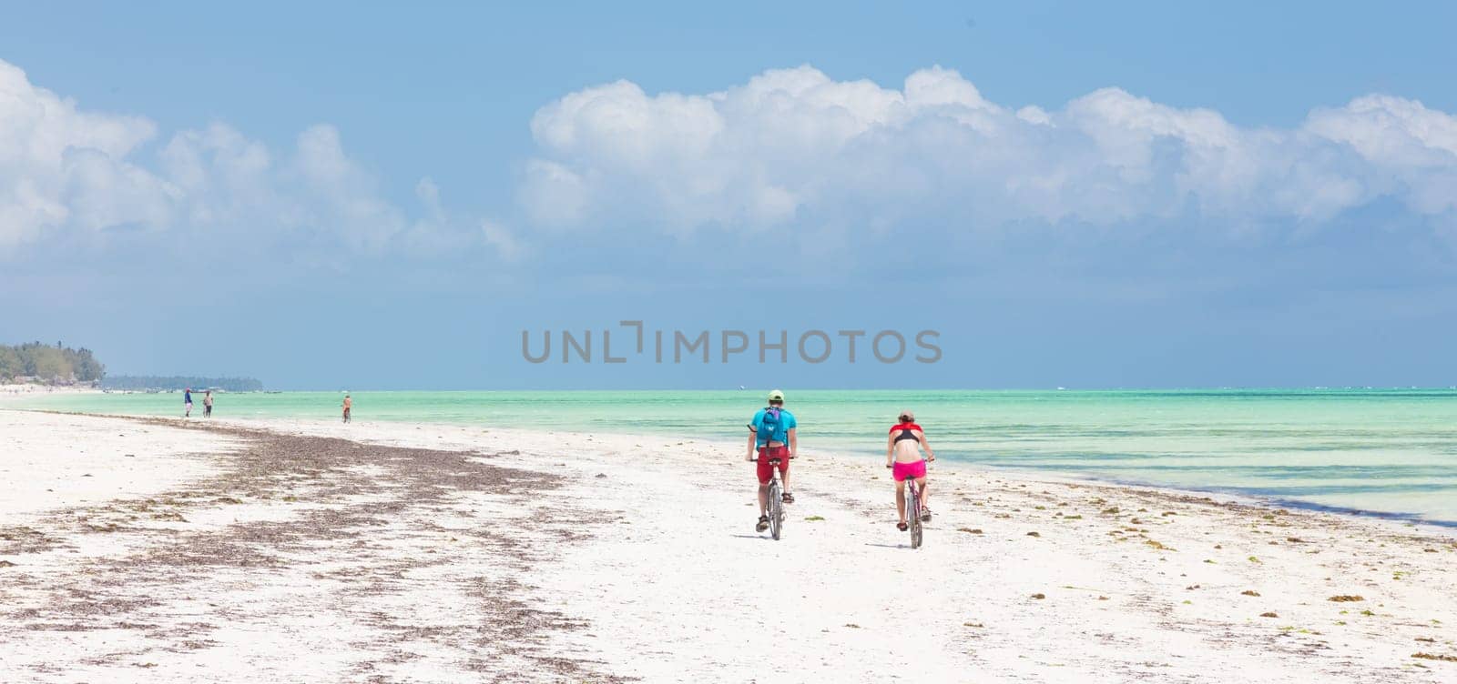 Active sporty tourist couple cycling down picture perfect white sand tropical beach of Paje village while being on vacations on Zanzibar, Tanzania.