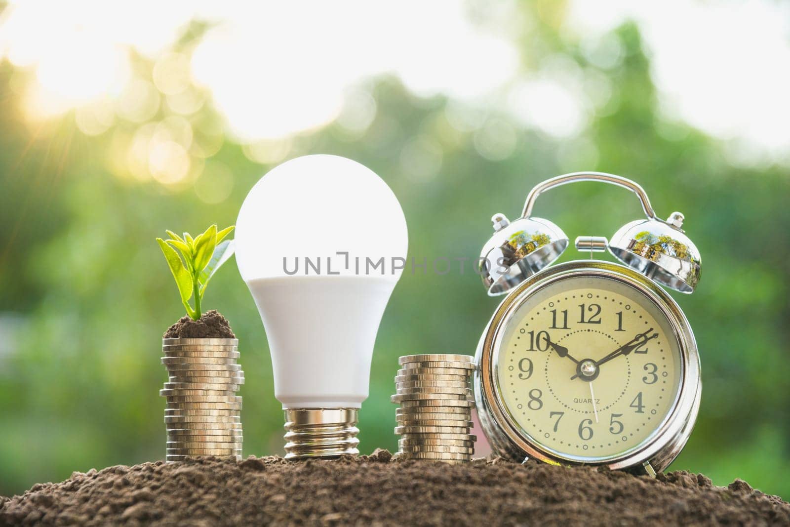 Energy saving light bulb and alarm clock on stacks of coins on nature background. Saving, accounting and financial concept.