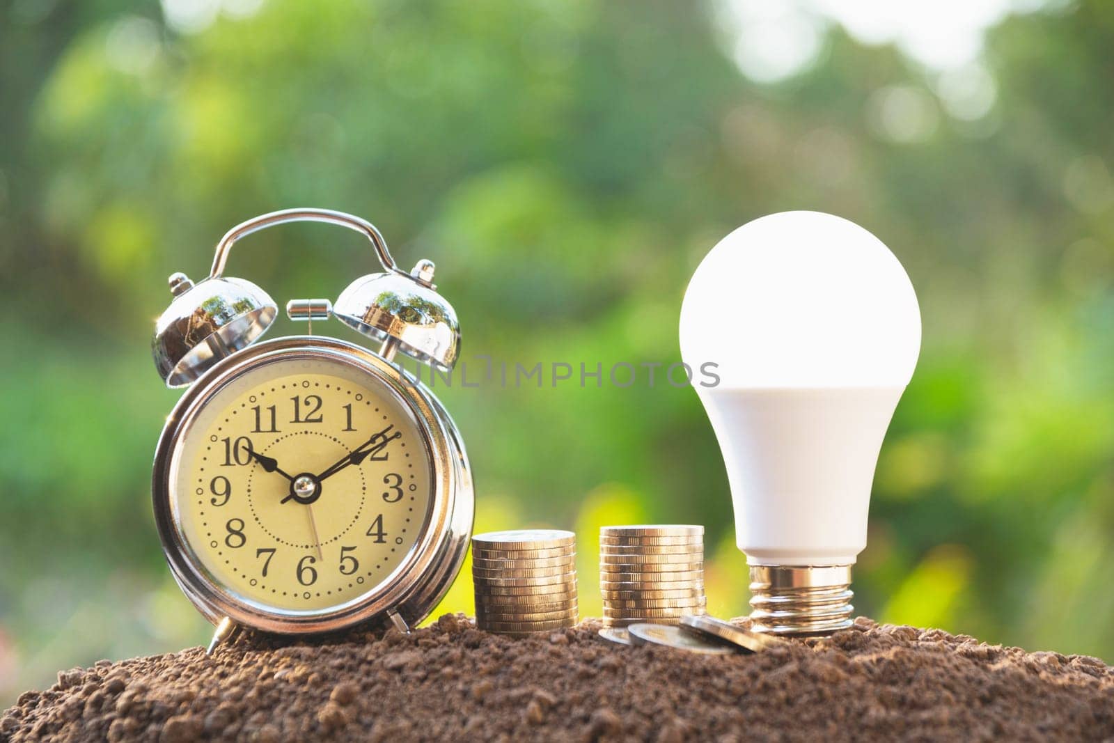 Energy saving light bulb and alarm clock on stacks of coins on nature background. Saving, accounting and financial concept.