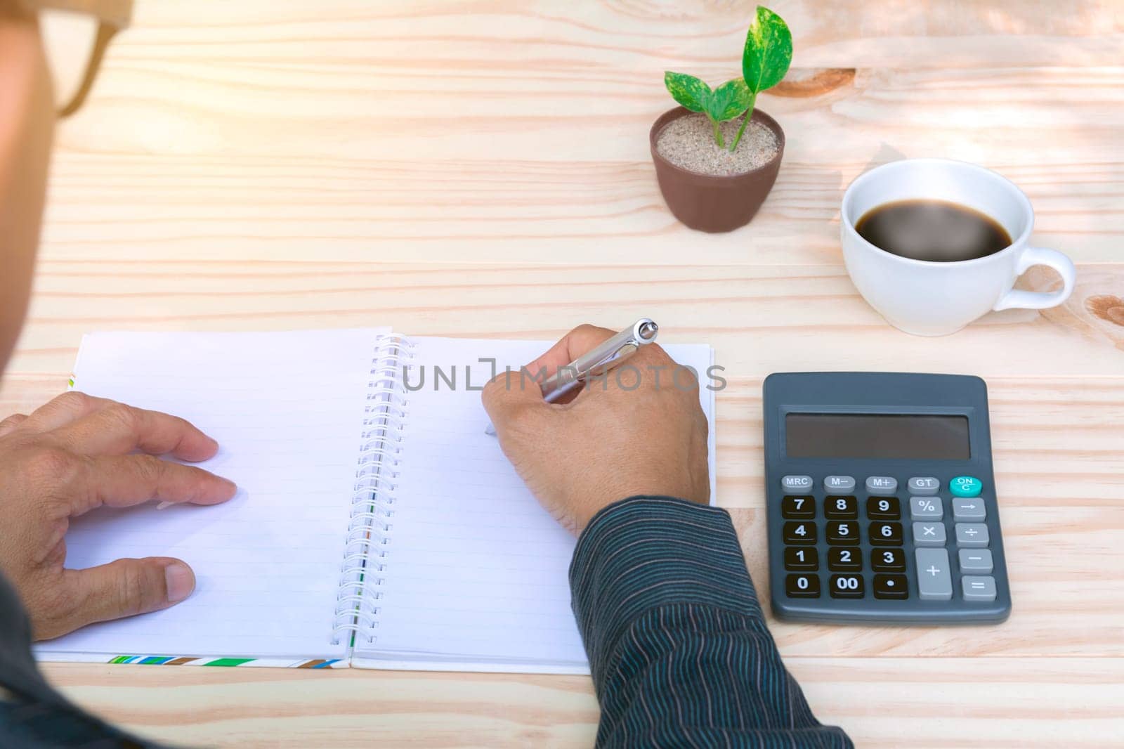 Person working and writing on notebook with calculator on wooden table background.