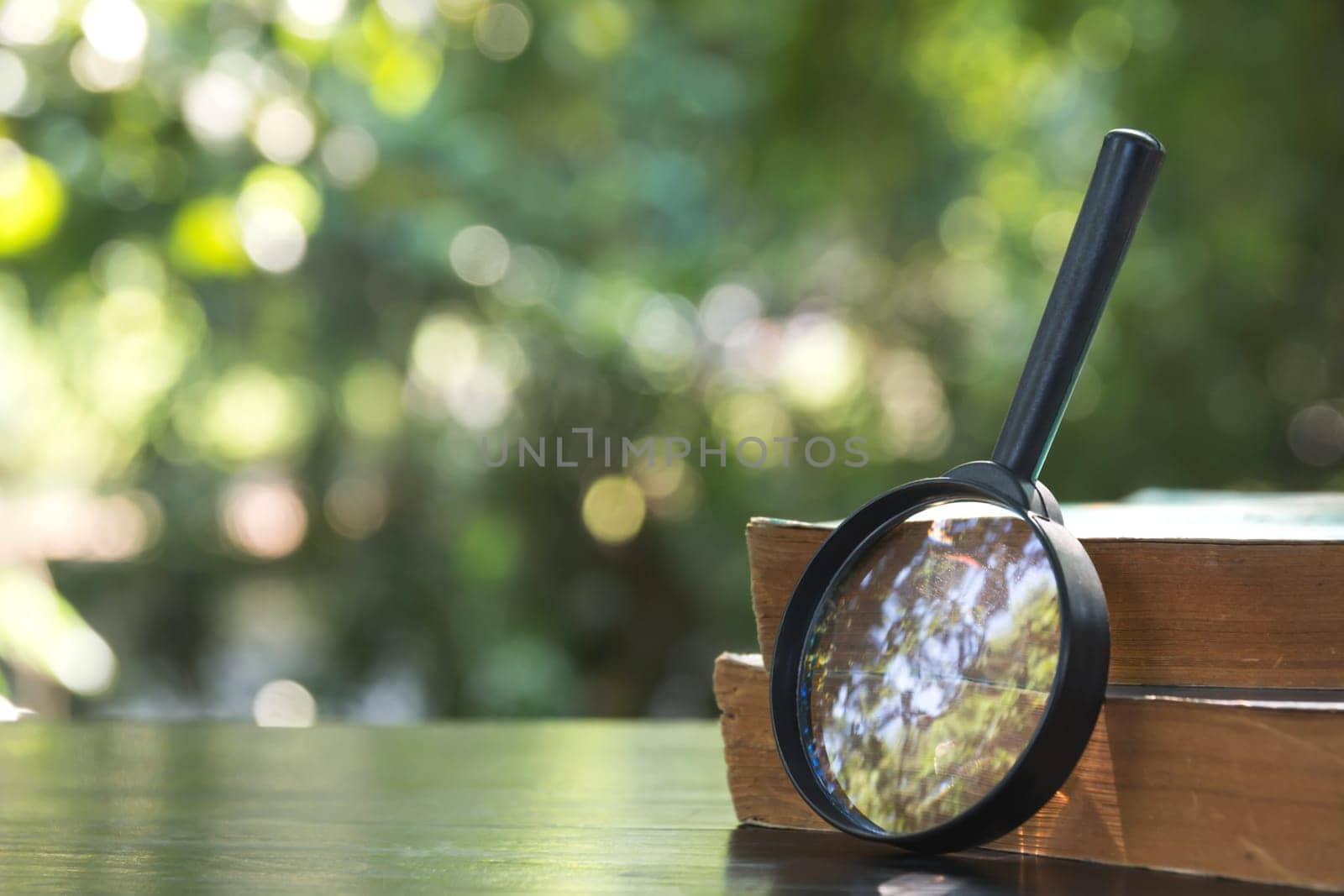 Close up of magnifying glass with old books on the wooden table and nature background.