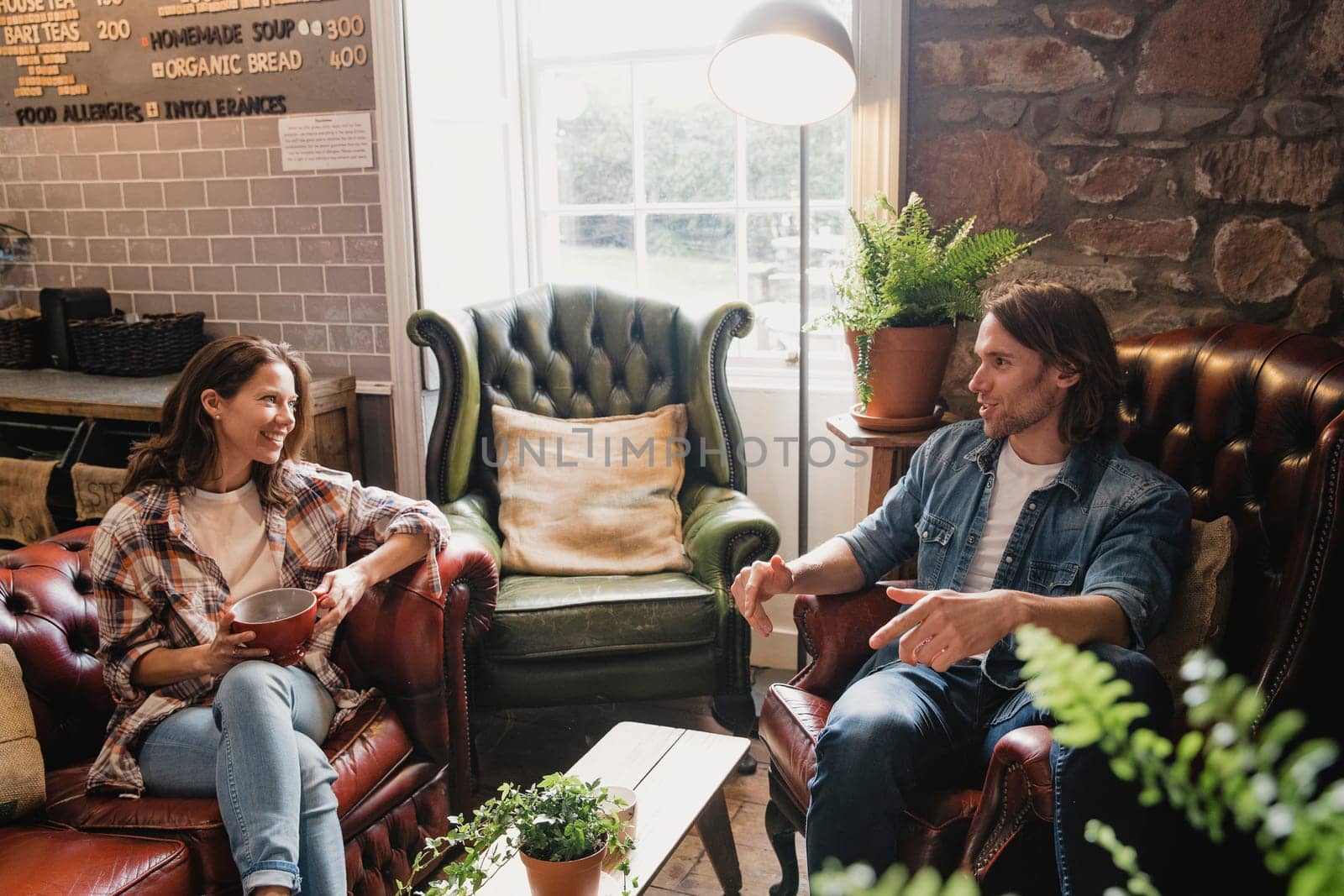 Mid-adult couple sitting in a small coffee shop relaxing. They are sitting on leather sofas and enjoying a coffee while talking.