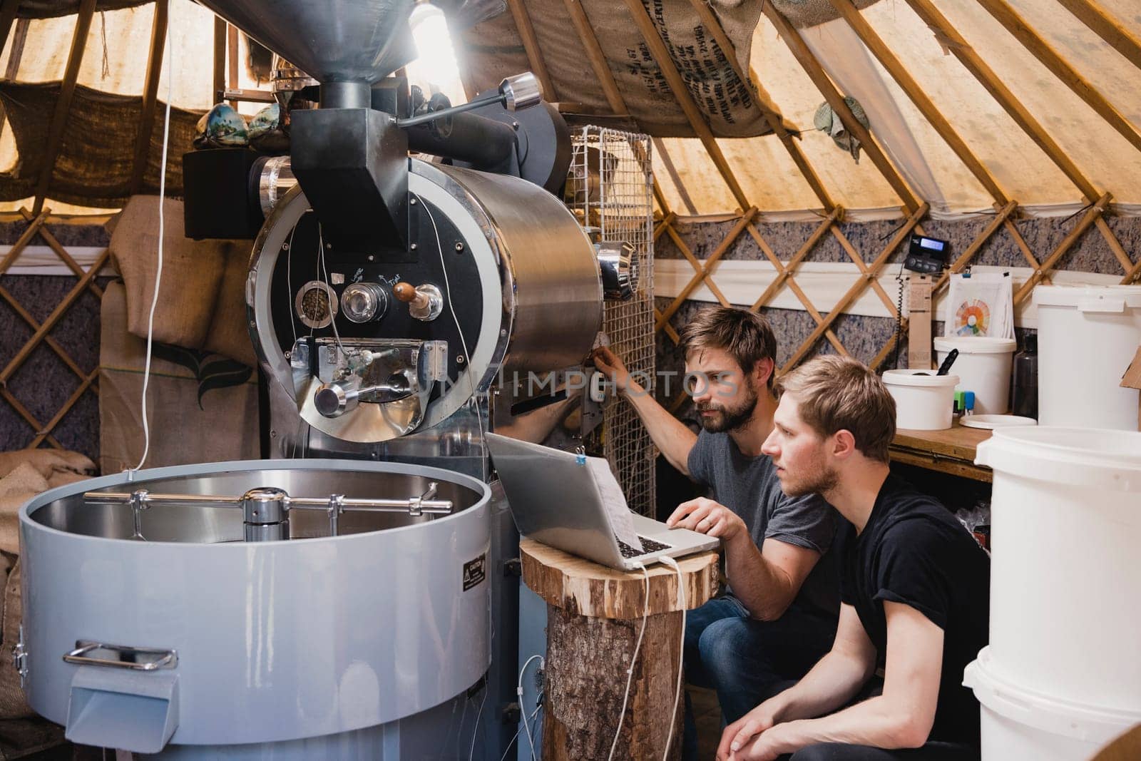 Two mid-adult men sitting in a room with a coffee bean roasting machine. They are looking at the laptop and fixing the machine, making sure it is working.