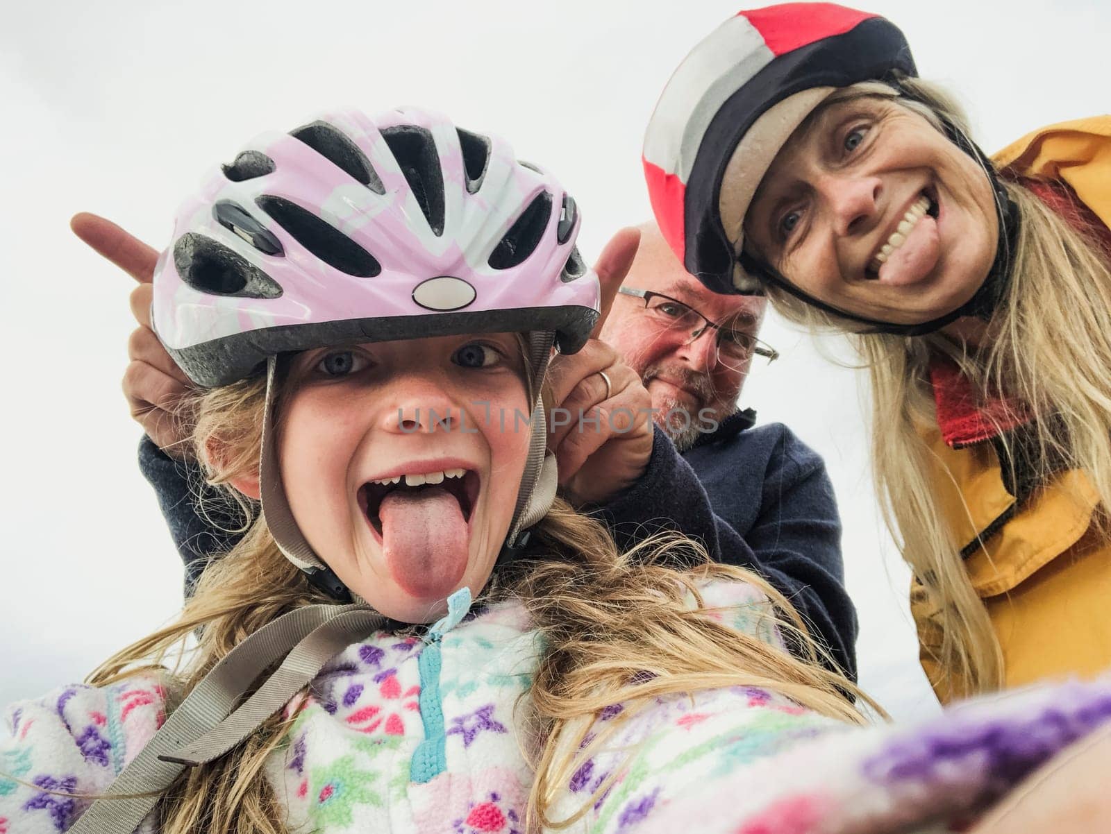 A close-up selfie of a happy family with one child, they are on an outdoor bikeride and pulling silly faces.