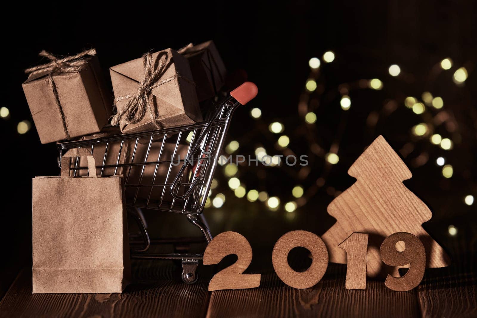 Wooden fir tree, gift boxes, shopping trolley and text 2019 from wooden figure on dark wooden background with LED light garland. Horizontal view. New Year and Christmas.