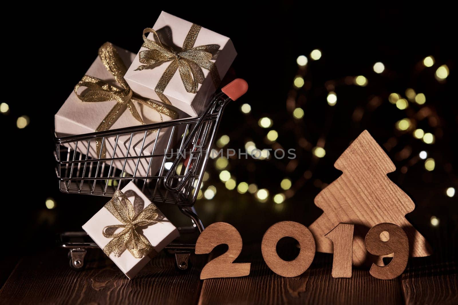 Wooden fir tree, gift boxes, shopping trolley and text 2019 from wooden figure on dark wooden background with LED light garland. Horizontal view. New Year and Christmas.