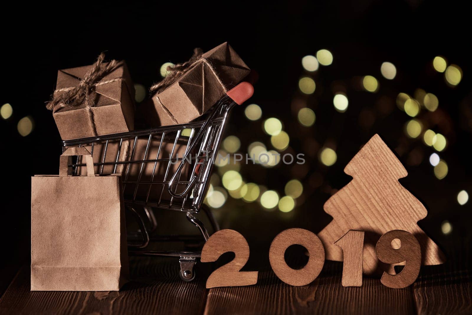 Wooden fir tree, gift boxes, shopping trolley and text 2019 from wooden figure on dark wooden background with LED light garland. Horizontal view. New Year and Christmas.
