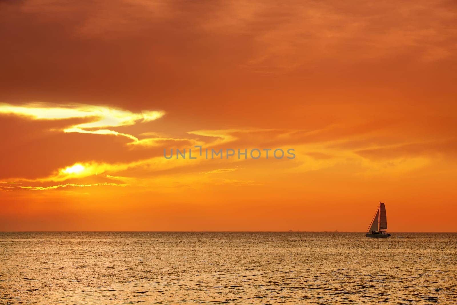 Orange color seascape image with shiny sea and sailboat over cloudy sky and sun during sunset in Cozumel, Mexico