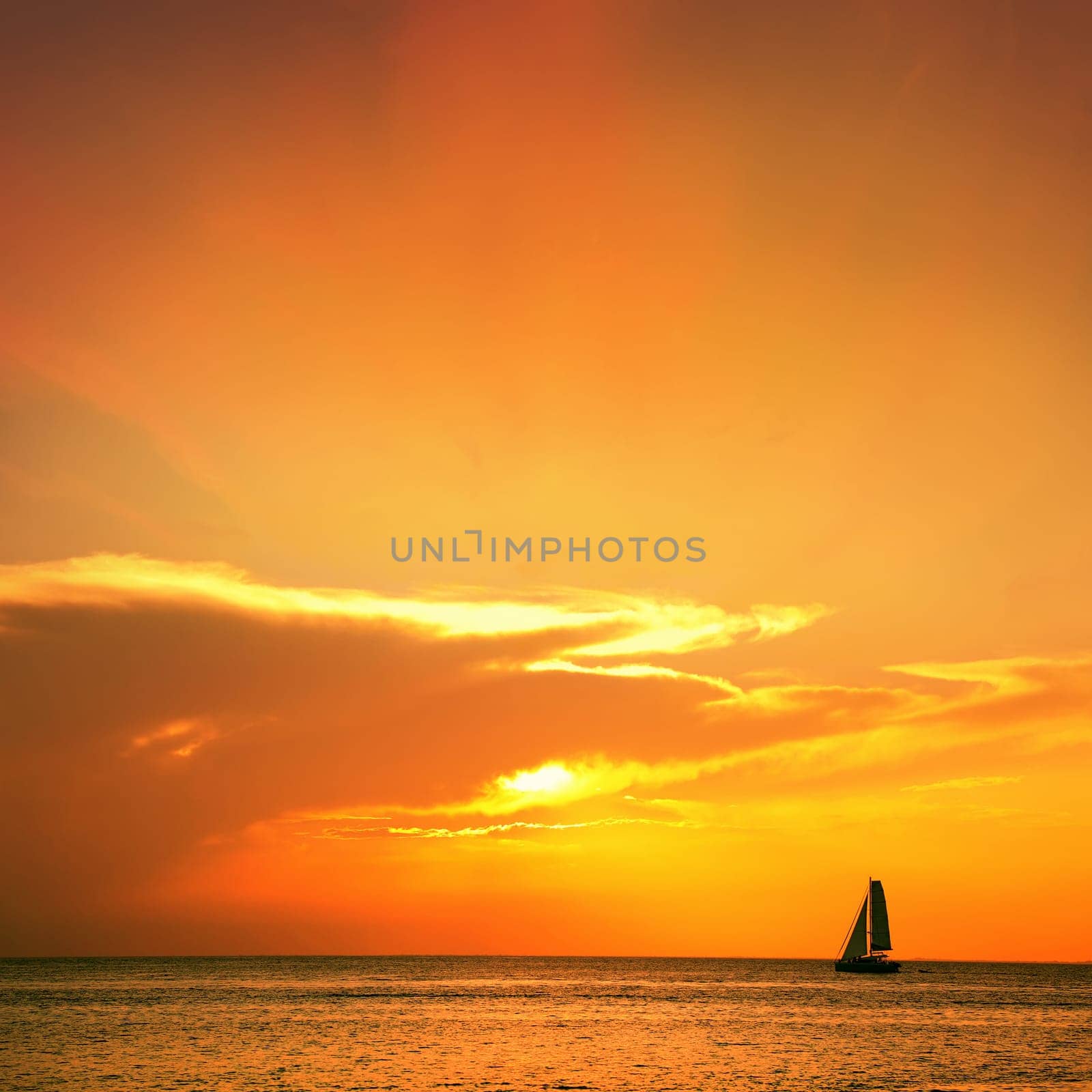 Orange color seascape image with shiny sea and sailboat over cloudy sky and sun during sunset in Cozumel, Mexico