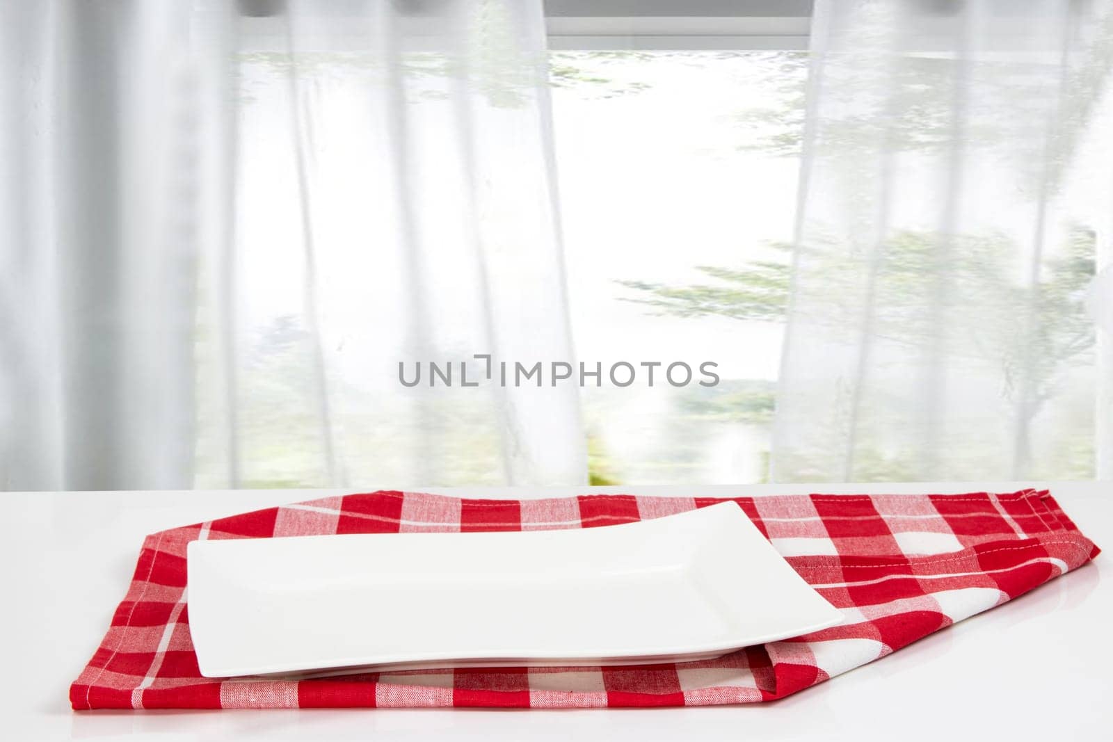 Empty plate background. Empty white plate on table with red napkin or tablecloth over blurred curtain window natural light green background. Template for your food and product display montage. Concept food. Copy space.