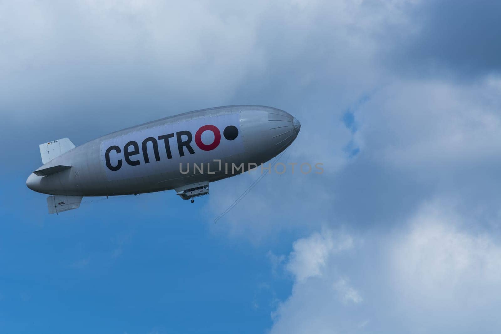 Heiligenhaus, Nrw, Deutschland - Juni 05, 2017:.Luftschiff, Zeppelin vor blauen Himmel mit dunklen Wolken und Werbebanner mit Aufschrift Centro O...HEILIGENHAUS, NRW, DEUTSCHLAND - JUNE 05, 2017:.Airship, zeppelin against blue sky with dark clouds and banners with label Centro O.