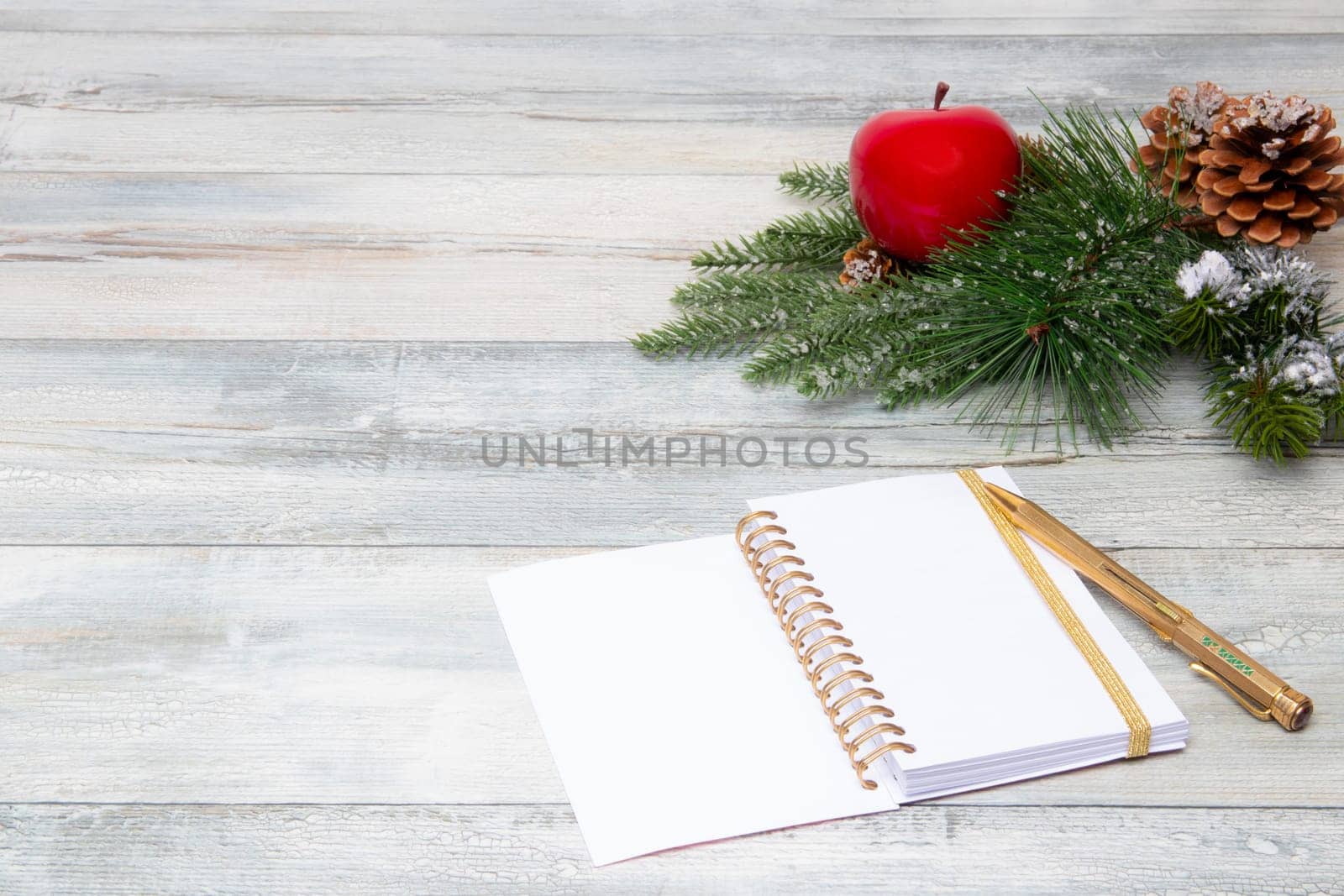 Christmas decoration background. Composition from sparkling christmas tree branches with a red apple and fir cones and a blank notebook with pen on a rustic wooden background. Top view copy space.