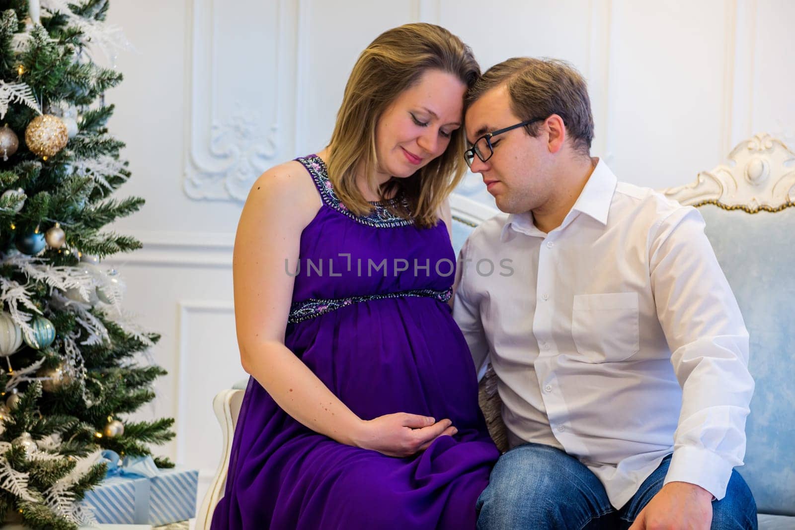 Pregnant woman in ultra violet dress with husband sitting on the sofa near christmas tree
