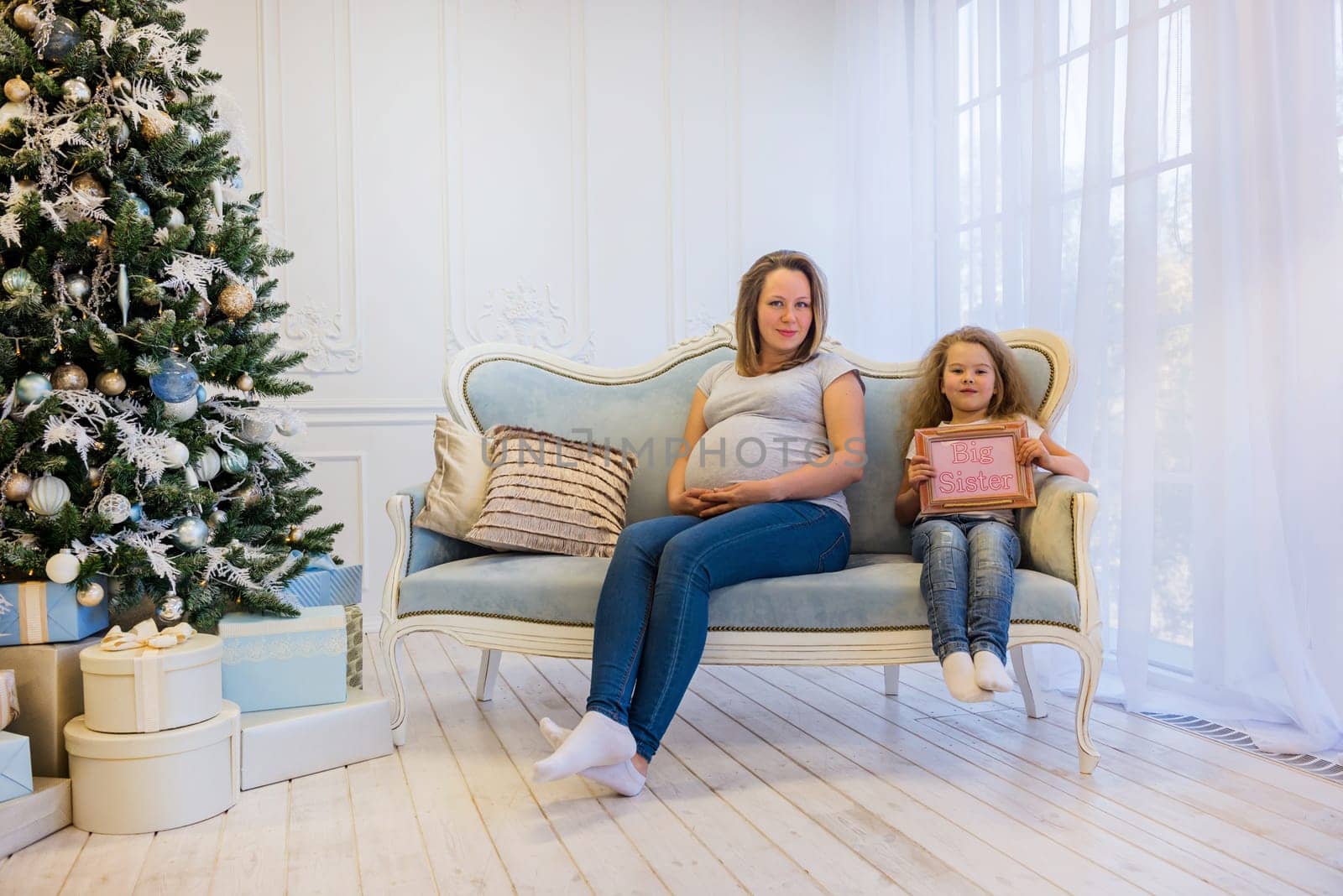 Beautiful pregnant woman sitting with daughter on the sofa near christmas tree