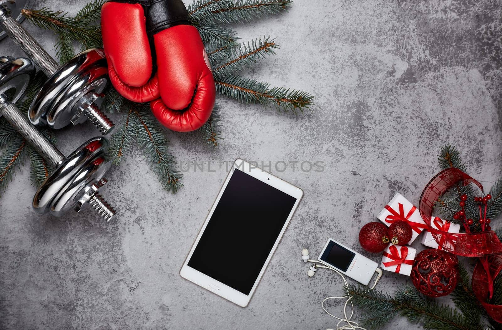 Dumbbells, boxing gloves, computer tablet, fir tree branches and Christmas decorations on a gray background.Top view with copy space. New Year and Christmas. Fitness, sport and healthy lifestyle concept.