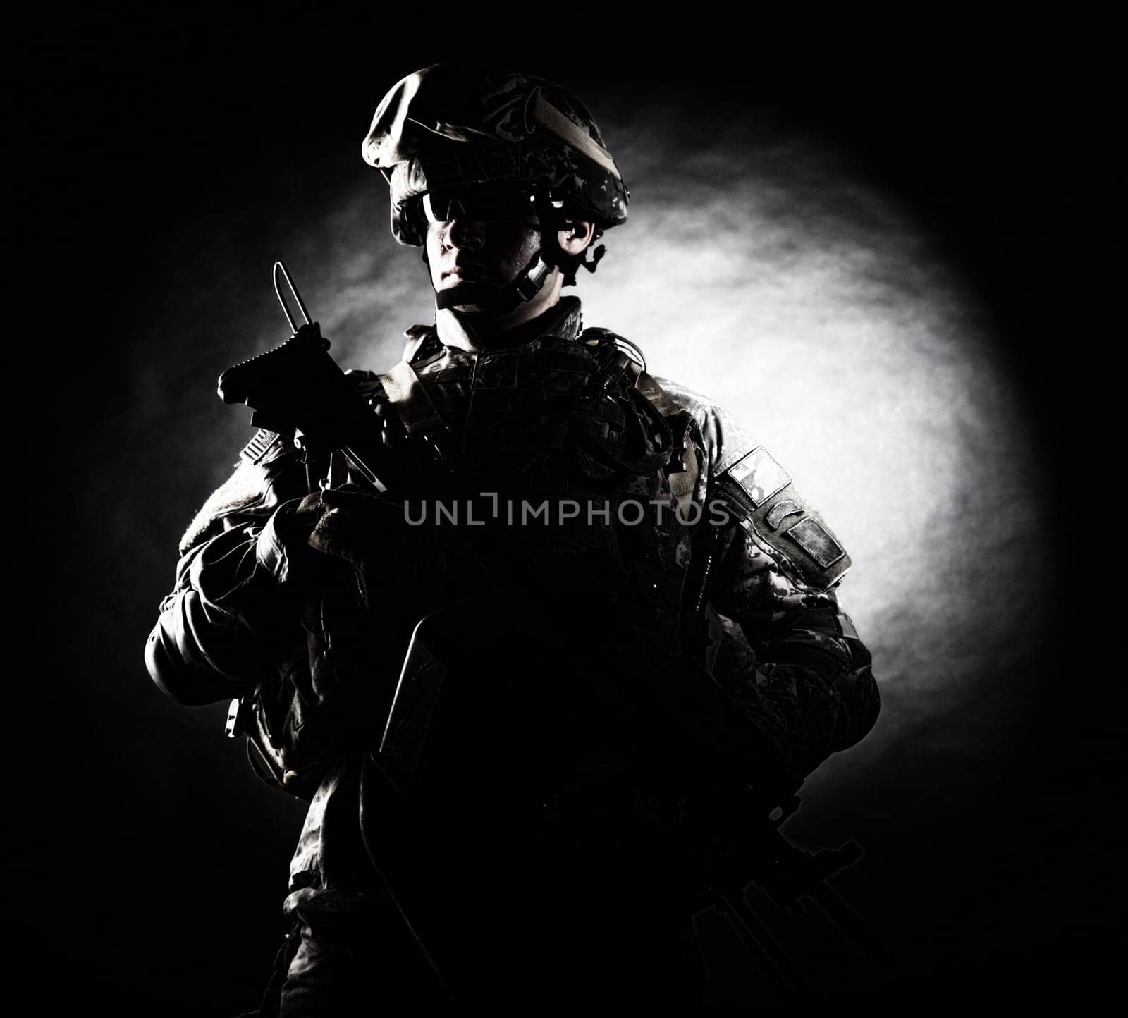 Low key studio portrait of private security service contractor, army infantry rifleman, US marine raider in helmet, sunglasses, camouflage uniform posing with weapon on black background with backlight