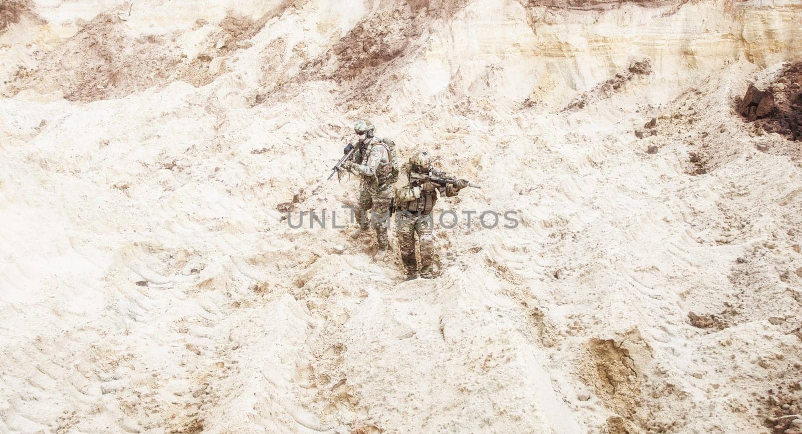 Two infantry soldiers in combat camo uniform, with tactical ammunition, carefully walking and aiming with assault rifles in unknown desert area. Military reconnaissance, scouting on enemy territory