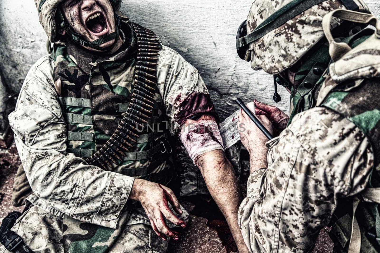 Marine wounded in shoulder, suffering of pain and screaming while receiving medical aid from comrade. Military medic apply pressure bandage to casualty, binding gunshot wound, trying stop bleeding