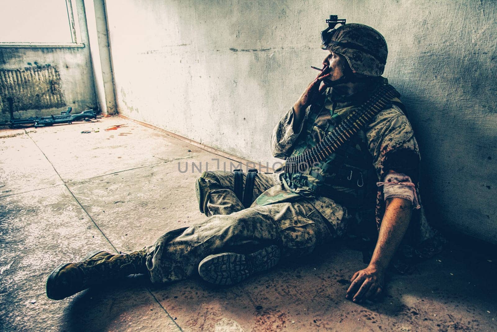 Soldier in combat uniform and ammo belt over chest, sitting on floor and smoking after being shoot in shoulder and put bandage on wounded hand, dropped weapon lying nearby with blood stains around