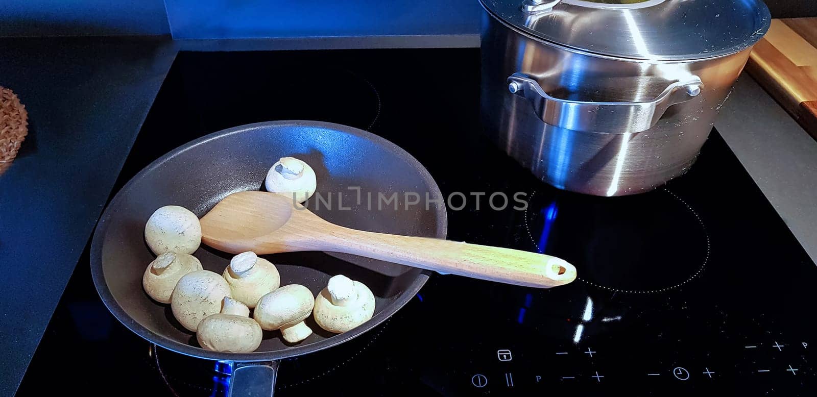 A silver pot with boiling noodles and a cast iron frying pan on a black ceramic hob