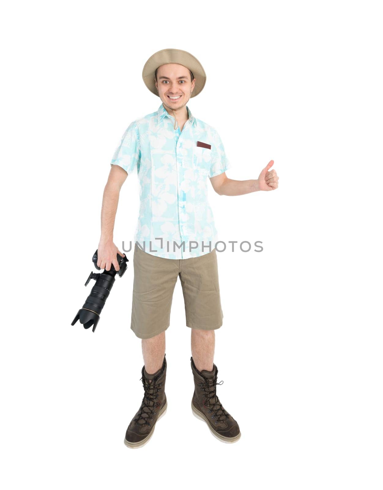 Happy man tourist in blue shirt, brown shorts and hat holds big dslr camera, giving thumbs up and smiles. Traveler going to photo tour. Isolated on white background.