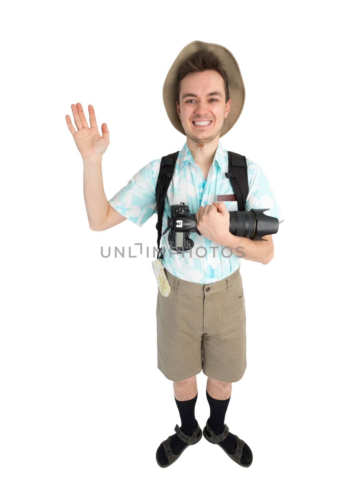 Happy man tourist in blue shirt, brown shorts and hat with backpack on his shoulders holding dslr camera. Traveler smiling and greeting someone by hand. Isolated on white background.
