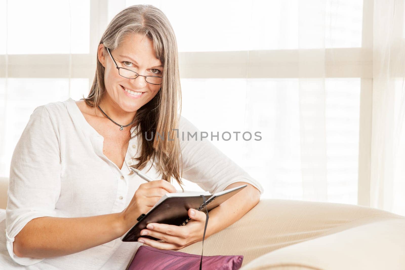 Mature woman is making notes in her diary sitting on a couch in her apartment