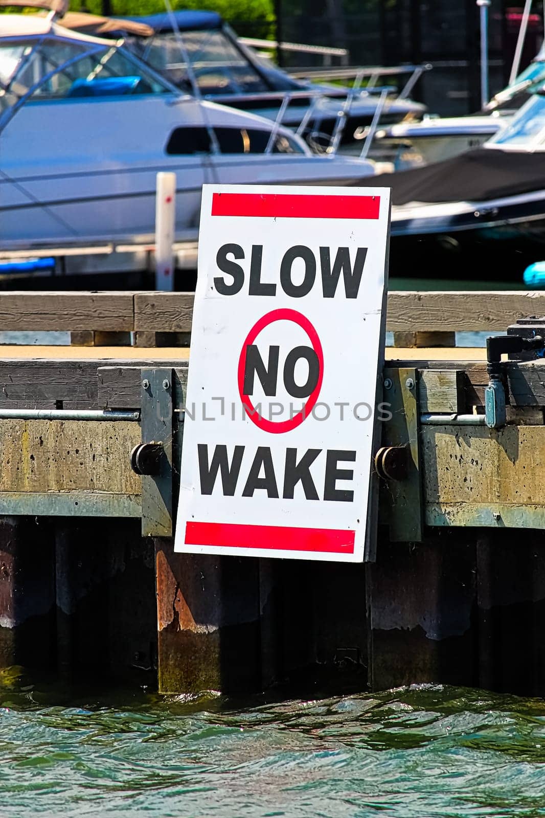 A slow no wake sign on a dock with boats in the background.