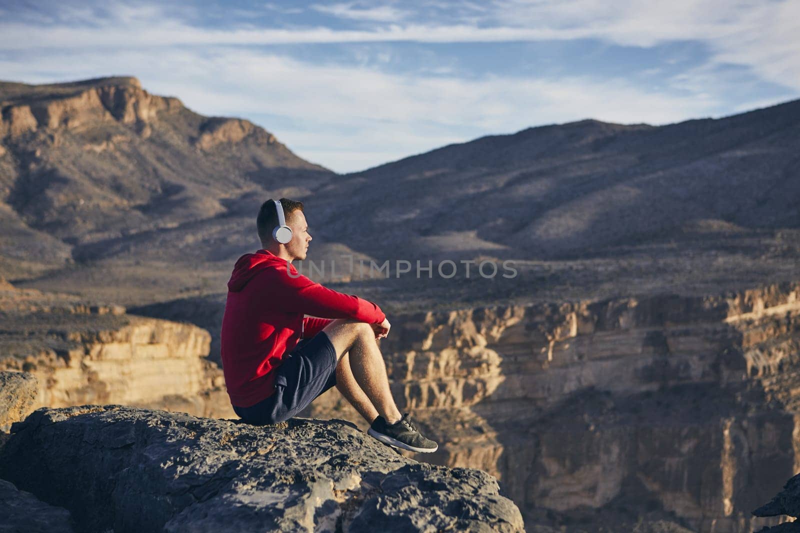 Relaxation in mountains. Young man with headphones sitting on the edge of cliff and listening music. Jebel Akhdar, Grand Canyon of Oman.