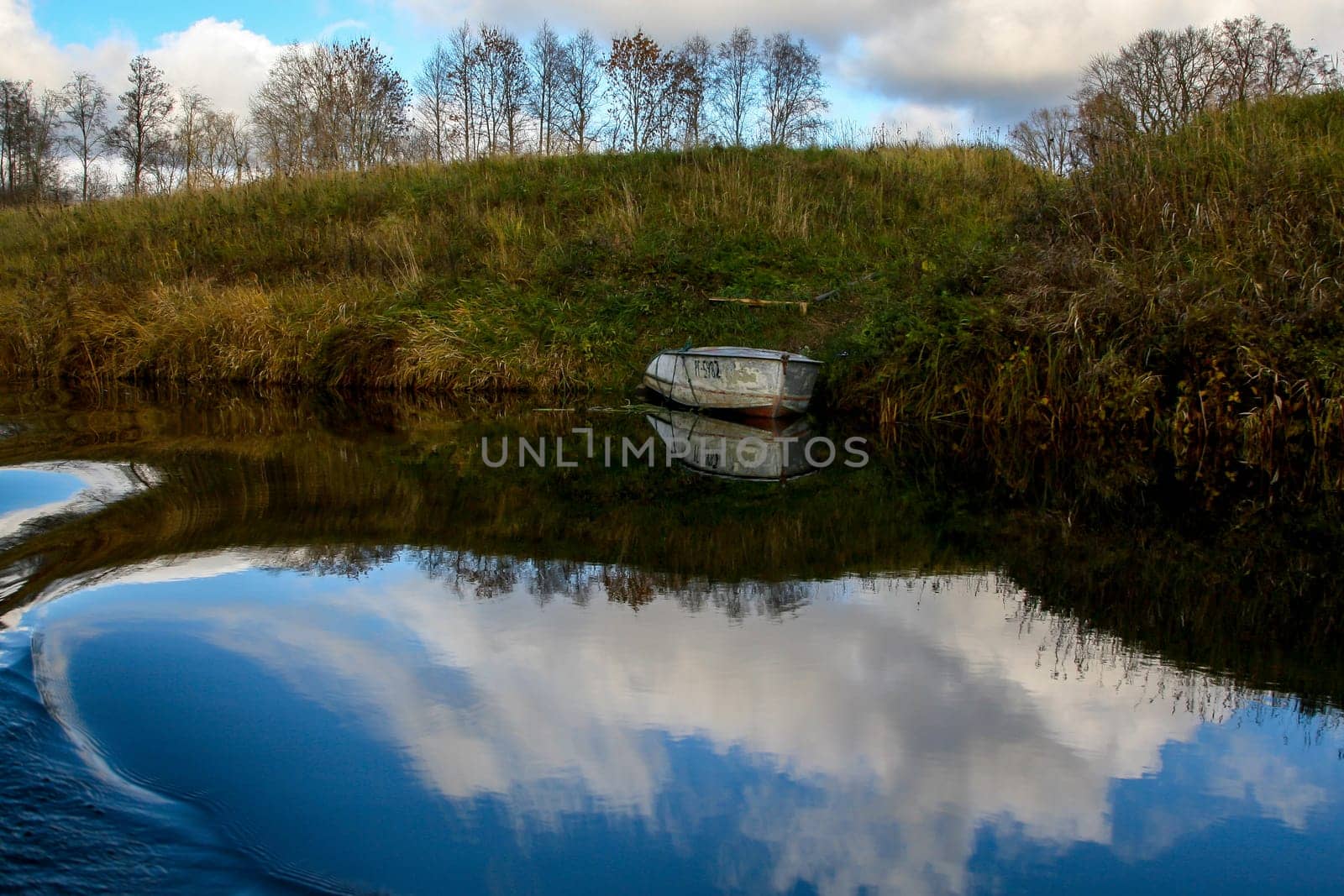 View on autumn landscape of river and boat in sunny day. Forest on river coast in autumn day. Reflection of coast, clouds  and boat in water. Autumn in Latvia. Autumn landscape with trees grass and boat in river.