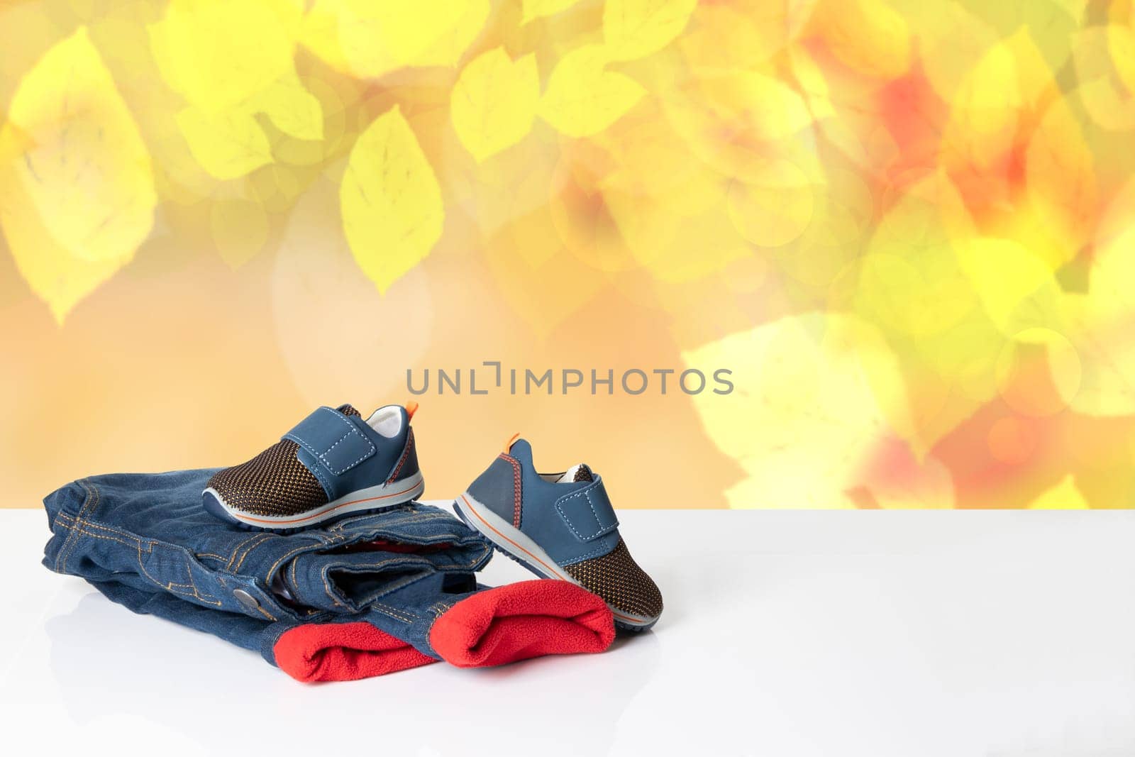 Colorful children jeans clothes.  Fashionable red lined thermojeans and a trendy pair of blue sneakers on bright table over abstract yellow autumn background. Autumn and winter jeans fashion. Space for your display product montage.