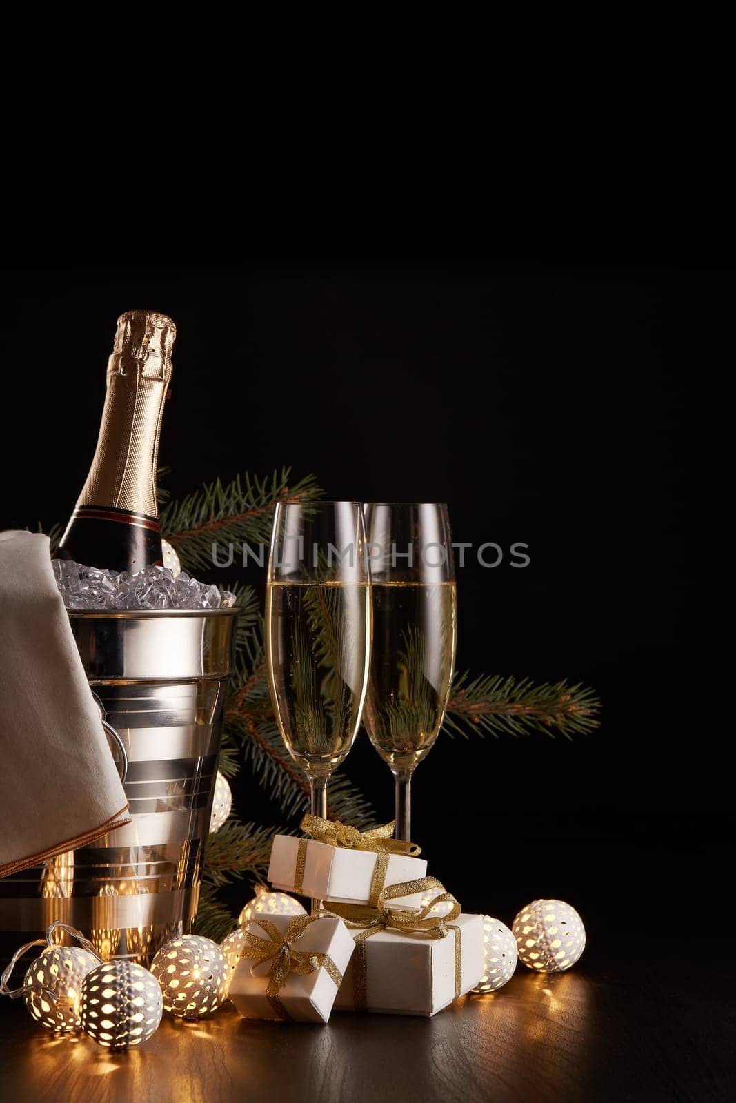 Still life with champagne bottle standing in a bucket with ice, two full champagne flutes, LED lights garland and Christmas ornaments on a black background. Christmas and New Year celebration