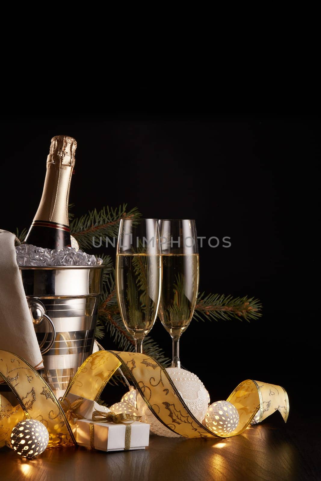 Still life with champagne bottle standing in a bucket with ice, two full champagne flutes, LED lights garland and Christmas ornaments on a black background. Christmas and New Year celebration