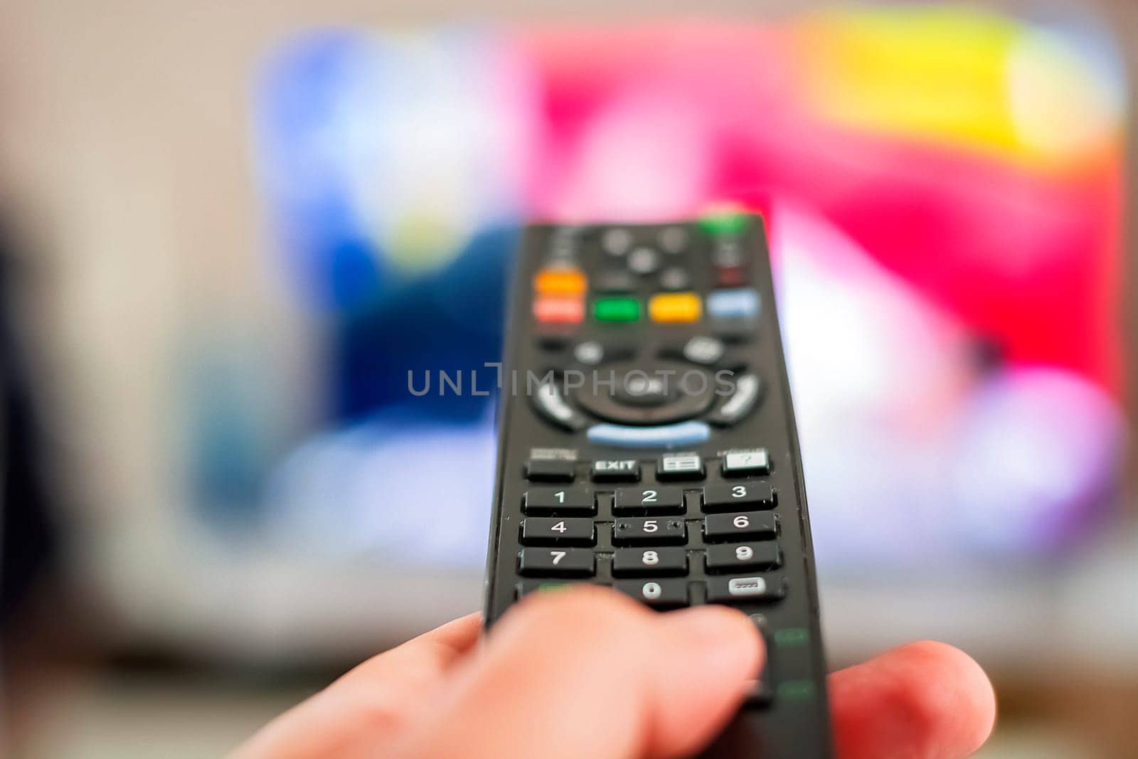 A hand pressing a remote control button to change channels in front of a blurry television screen